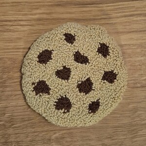 May include: A brown and beige crocheted cookie with chocolate chips. The cookie is round and has a textured surface.