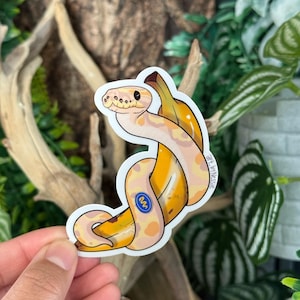 May include: A sticker featuring a cartoon snake wrapped around a banana. The snake is light tan with darker spots, and the banana is yellow with brown spots. The sticker has the text "art.by.nessie" and a blue logo.