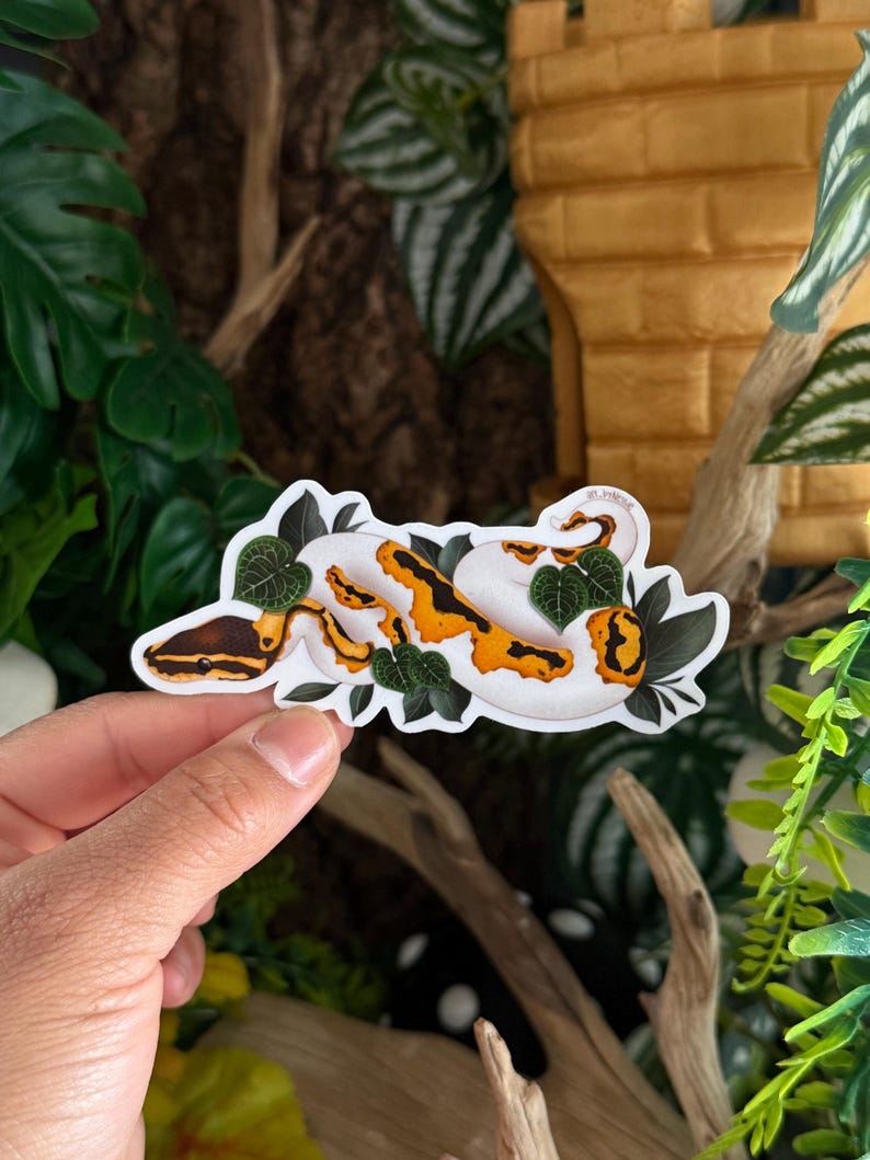 May include: A sticker depicting a white, orange, and brown snake design, with green leaves. The snake is curled up, with its head and tail visible. The sticker is held by a hand against a backdrop of greenery and a tan structure.