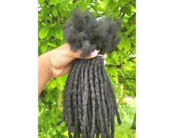 Op de afbeelding: Een bundel zwarte dreadlocks van echt haar, vastgehouden in een hand tegen een groene, bladerrijke achtergrond.