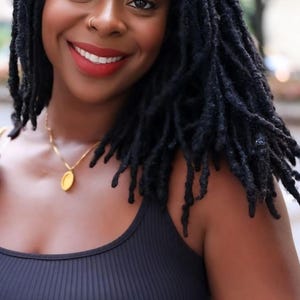 May include: A smiling person with black dreadlocks, wearing a black tank top and a gold pendant necklace. The image includes the text "Afrikan Locs" with a logo featuring the colors of the Pan-African flag.