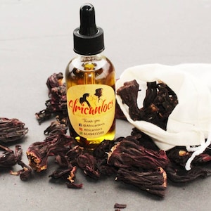 May include: A glass bottle with a dropper filled with a golden liquid. The bottle has a label that says "Africanlocs" with social media handles and a phone number. The bottle is surrounded by dried red hibiscus flowers.