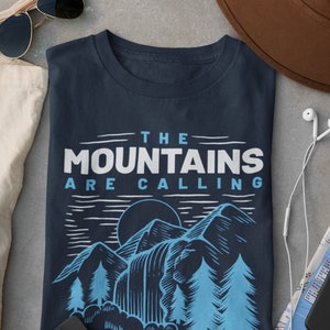Puede incluir: Una camiseta azul marino con un diseño gráfico de montañas, árboles y una cascada. El texto "THE MOUNTAINS ARE CALLING" está impreso en blanco en la camiseta.
