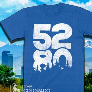 May include: A royal blue t-shirt with a white distressed graphic of the numbers 5280 with a silhouette of trees behind the numbers. The text "THE COLORADO COAST" is printed in white below the graphic.