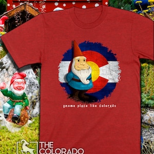 Pode incluir: Camiseta vermelha com um gráfico de um gnomo em pé em frente a uma bandeira do Colorado desbotada. O texto "gnome place like colorado" é impresso abaixo do gráfico. O logotipo "THE COLORADO COAST" é impresso na parte inferior da camiseta.