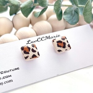 May include: A pair of square stud earrings with a leopard print design. The earrings are displayed on a white card with the text "Handmade Clay Couture" and the Etsy shop URL. The background includes wooden beads and greenery.