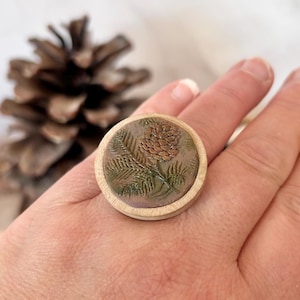 May include: A round ring with a light wood frame. The ring's center features a detailed design of a pinecone and fern leaves in shades of brown and green. The ring is worn on a hand.