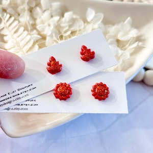 May include: Red glittery stud earrings in two designs: a Christmas tree and a flower. The earrings are displayed on white cards. The background includes white flowers and a white dish.