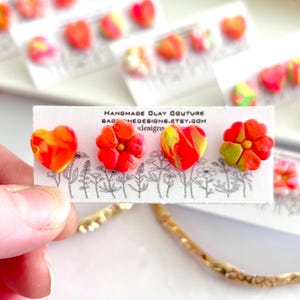 May include: Four stud earrings made of clay attached to a white display card. The earrings feature two heart shapes and two flower shapes in marbled red, orange, and yellow colors. The card displays the text HANDMADE CLAY COUTURE.