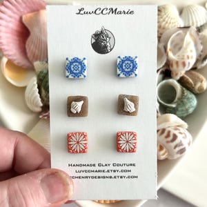 May include: A card featuring six pairs of square stud earrings. The earrings have various designs: blue and white tile patterns, seashell accents on brown squares, and red and white floral patterns. The card reads "LuvCCMarie" and "Handmade Clay Couture".