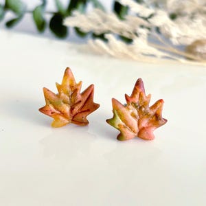 May include: A pair of leaf-shaped stud earrings. The earrings are in the shape of maple leaves, with a mix of orange, yellow, and green colors. The earrings are on a white surface.