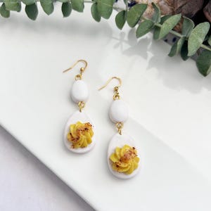May include: A pair of gold dangle earrings with white and yellow clay egg-shaped charms. The yellow charms have a swirl design and resemble deviled eggs.