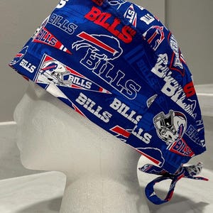 May include: A blue and red patterned surgical scrub cap featuring the text "BILLS" and the Buffalo Bills logo. The cap is tied at the back with matching fabric ties. The design includes football helmets and team flags.