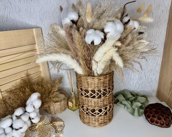 May include: A wicker vase with a woven pattern holds a bouquet of dried pampas grass and cotton stems. The vase is on a white surface with a wooden background.