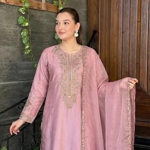 May include: A light pink embroidered salwar kameez set. The outfit includes a tunic, pants, and a sheer scarf. The tunic and pants feature intricate gold embroidery. The outfit is worn with matching shoes.