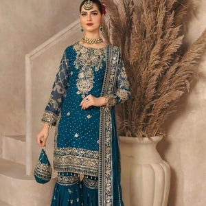 May include: A teal blue three-piece ensemble featuring a long-sleeved tunic, flared trousers, and a matching dupatta. The garments are heavily embellished with intricate gold embroidery, sequins, and beadwork. A small matching drawstring pouch is included.