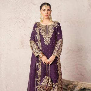 May include: A formal purple ensemble with intricate gold embroidery. The outfit includes a long-sleeved tunic, matching pants, and a small, embellished purse. The design features detailed patterns and a flowing silhouette, suitable for special occasions.