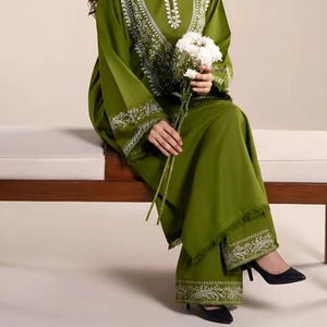 May include: Olive green two-piece outfit with white embroidered detailing. The long-sleeved top and wide-leg pants feature intricate patterns around the neckline, cuffs, and hem. The outfit is paired with black heels and a bouquet of white flowers.
