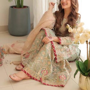 May include: A woman wearing a mint green and gold embroidered outfit with a matching dupatta. The outfit features intricate embroidery and red trim. She is sitting near a window, holding a flower, and wearing pink flats.