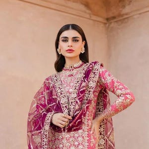 May include: A vibrant pink long-sleeved tunic and matching dupatta featuring intricate gold embroidery, sequin detailing, and a high neckline. The ensemble includes a coordinating deep purple drawstring pouch with gold embellishments.