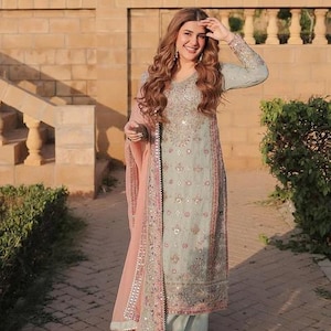 K&ouml;nnte beinhalten: Ein helles salbeigr&uuml;nes Set aus lang&auml;rmeliger Tunika und weit geschnittener Hose mit aufwendigen Silberstickereien, Pailletten und Spiegelapplikationen. Dazu geh&ouml;rt ein transparenter pfirsichfarbener Dupatta mit passenden Bord&uuml;ren.
