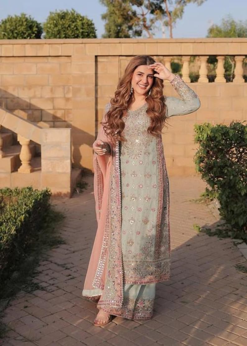 K&ouml;nnte beinhalten: Ein helles salbeigr&uuml;nes Set aus lang&auml;rmeliger Tunika und weit geschnittener Hose mit aufwendigen Silberstickereien, Pailletten und Spiegelapplikationen. Dazu geh&ouml;rt ein transparenter pfirsichfarbener Dupatta mit passenden Bord&uuml;ren.
