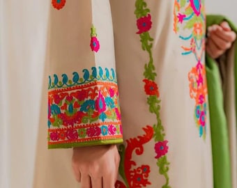 May include: Beige long-sleeved garment with vibrant floral embroidery in pink, orange, green, and blue. The sleeve cuff features a detailed border. A green shawl is held in the background. The garment is likely part of a traditional outfit.