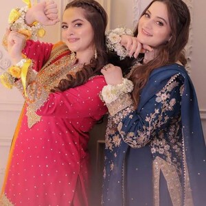May include: Two women in ornate, embroidered traditional dresses. One wears a red dress with gold accents, the other a navy blue dress with gold embroidery. Both have floral wrist and arm adornments.