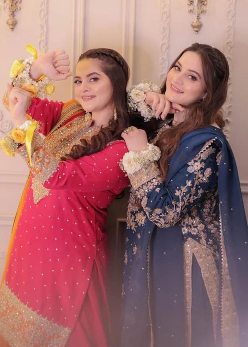 May include: Two women in ornate, embroidered traditional dresses. One wears a red dress with gold accents, the other a navy blue dress with gold embroidery. Both have floral wrist and arm adornments.