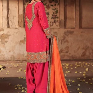 May include: A vibrant pink salwar kameez ensemble with intricate gold embroidery on the neckline, cuffs, and hem. The outfit includes a matching dupatta in orange with gold trim. The traditional South Asian attire is suitable for festive occasions.
