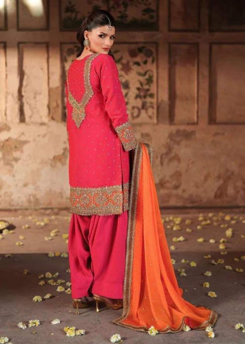 May include: A vibrant pink salwar kameez ensemble with intricate gold embroidery on the neckline, cuffs, and hem. The outfit includes a matching dupatta in orange with gold trim. The traditional South Asian attire is suitable for festive occasions.