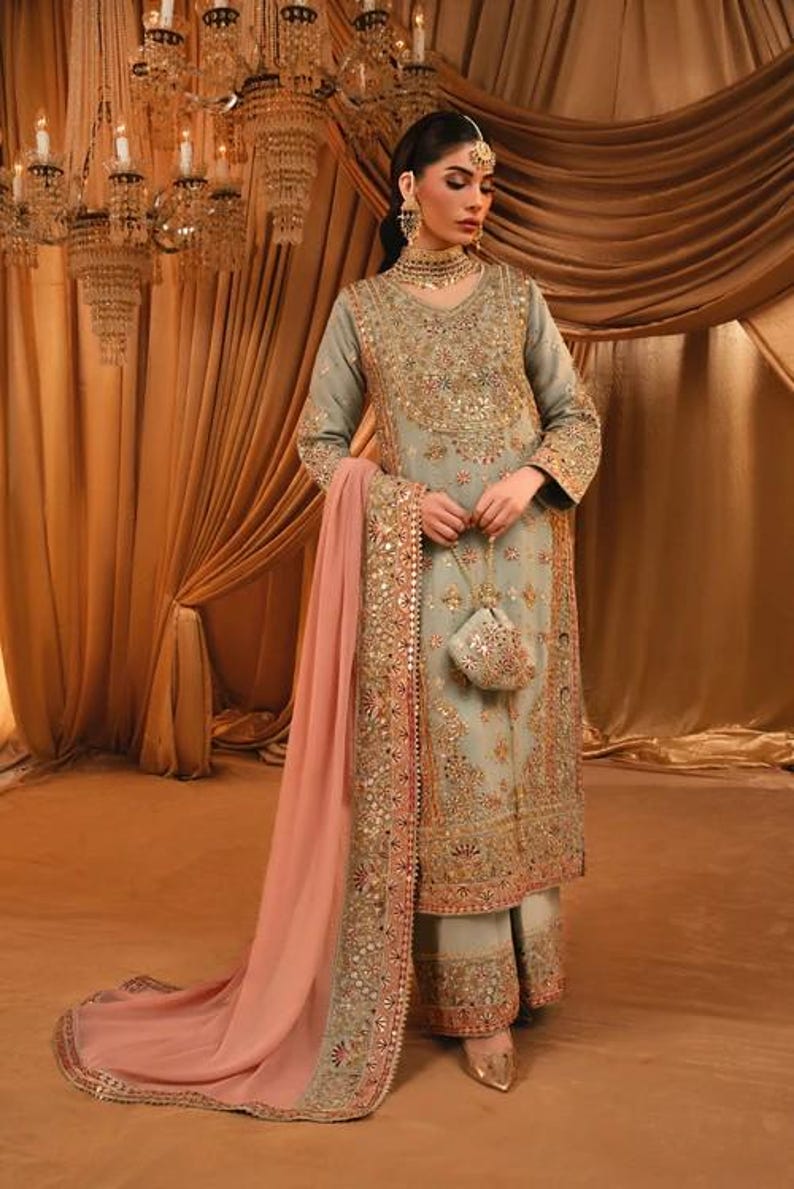 K&ouml;nnte beinhalten: Ein salbeigr&uuml;nes Set aus lang&auml;rmeliger Tunika und weiter Hose mit aufwendigen Goldstickereien, Pailletten und Perlenarbeiten. Das Outfit enth&auml;lt einen passenden salbeigr&uuml;nen Dupatta mit breitem Goldrand und einen kleinen Beutel.