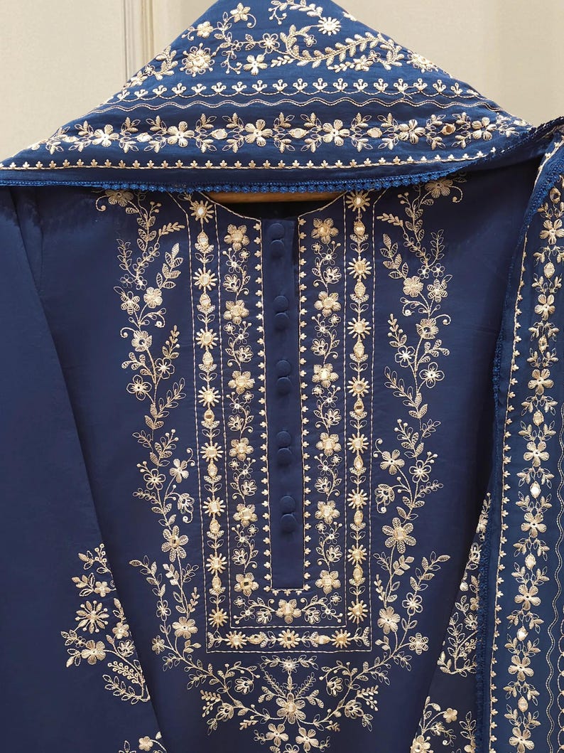 May include: A navy blue embroidered outfit featuring intricate gold floral patterns. The ensemble includes a tunic with a button-down front and a matching scarf, all adorned with detailed embroidery. The design is elegant and suitable for special occasions.