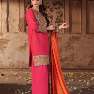 May include: A vibrant pink ensemble featuring a long-sleeved tunic and matching wide-leg pants. The tunic is adorned with gold embroidery on the sleeves and hem. An orange dupatta with gold trim drapes elegantly. The outfit is suitable for special occasions.