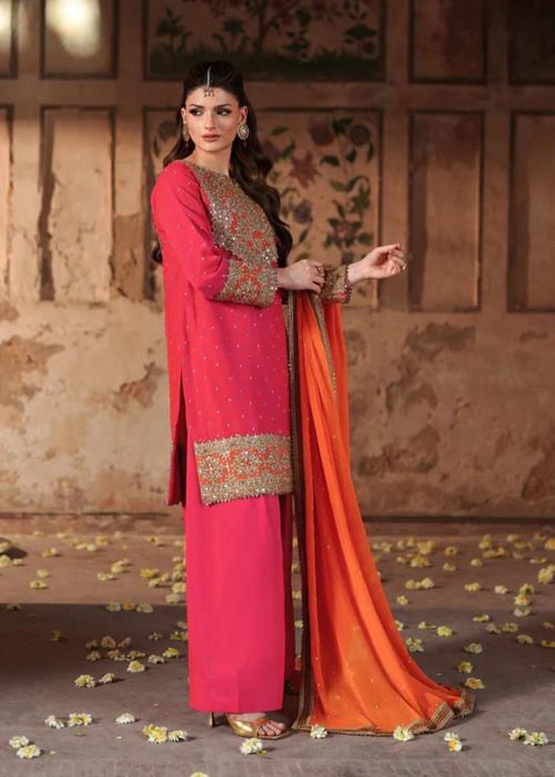 May include: A vibrant pink ensemble featuring a long-sleeved tunic and matching wide-leg pants. The tunic is adorned with gold embroidery on the sleeves and hem. An orange dupatta with gold trim drapes elegantly. The outfit is suitable for special occasions.
