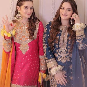 May include: Two women in traditional South Asian attire. One wears a vibrant pink outfit with gold embroidery and an orange scarf. The other is in a navy blue ensemble with gold detailing. Both outfits feature intricate patterns and embellishments.