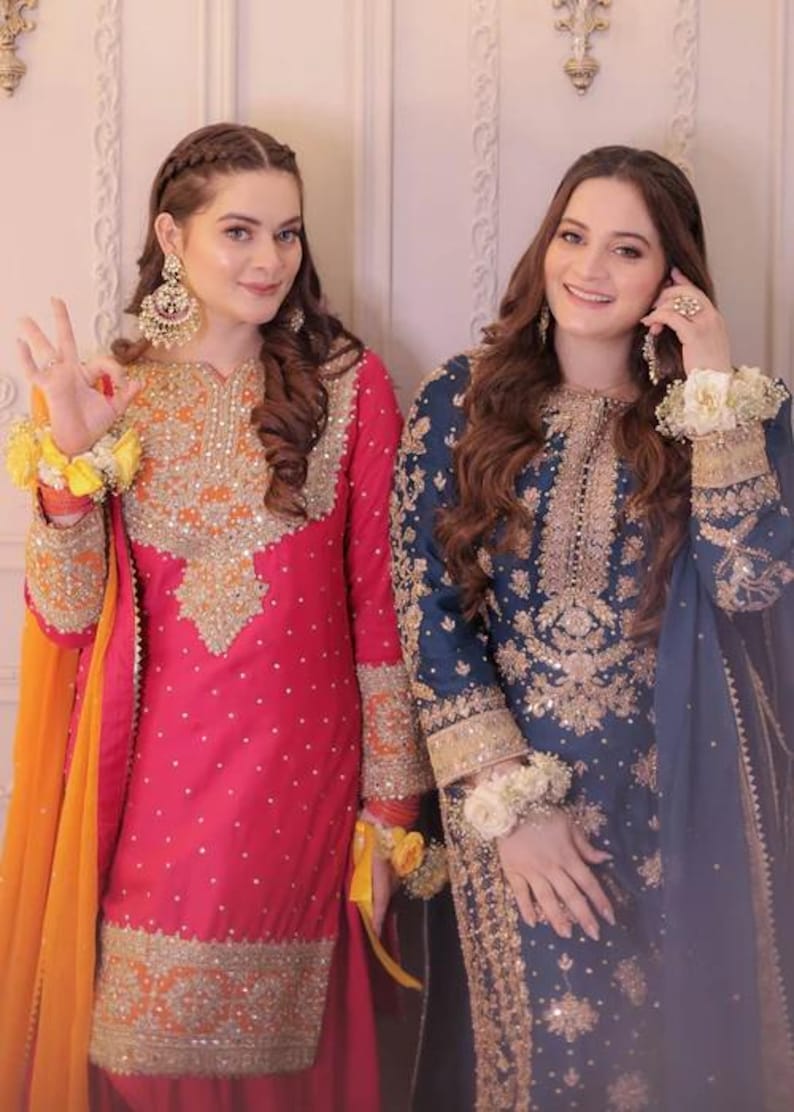 May include: Two women in traditional South Asian attire. One wears a vibrant pink outfit with gold embroidery and an orange scarf. The other is in a navy blue ensemble with gold detailing. Both outfits feature intricate patterns and embellishments.