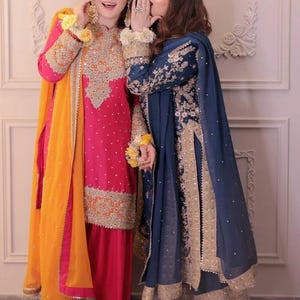 May include: Two women wearing traditional South Asian formal attire. One is in a vibrant pink outfit with gold embroidery and an orange scarf. The other is in a navy blue outfit with gold accents. Both outfits feature intricate detailing.