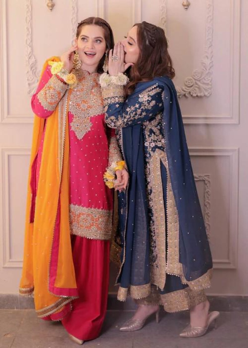 May include: Two women wearing traditional South Asian formal attire. One is in a vibrant pink outfit with gold embroidery and an orange scarf. The other is in a navy blue outfit with gold accents. Both outfits feature intricate detailing.