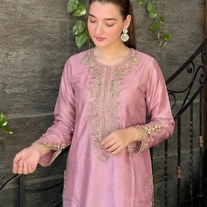 May include: A light pink, long-sleeved tunic and pants set. The tunic features intricate gold embroidery around the neckline, down the front, and on the cuffs. The outfit is made of a shiny, silky material, and is suitable for formal occasions.