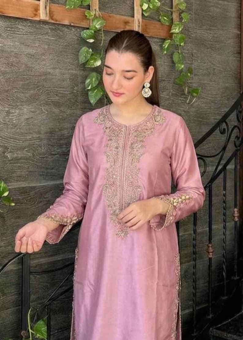 May include: A light pink, long-sleeved tunic and pants set. The tunic features intricate gold embroidery around the neckline, down the front, and on the cuffs. The outfit is made of a shiny, silky material, and is suitable for formal occasions.