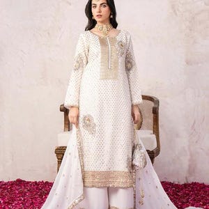 May include: A white embroidered salwar kameez with gold accents. The long-sleeved tunic features intricate gold embroidery and a matching dupatta. The outfit includes wide-leg pants and a small, decorative purse. The ensemble is suitable for special occasions.