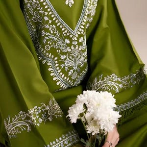 May include: Olive green tunic with intricate white floral embroidery around the neckline and sleeves. The long-sleeved garment features a V-neck design and is paired with a bouquet of white flowers.