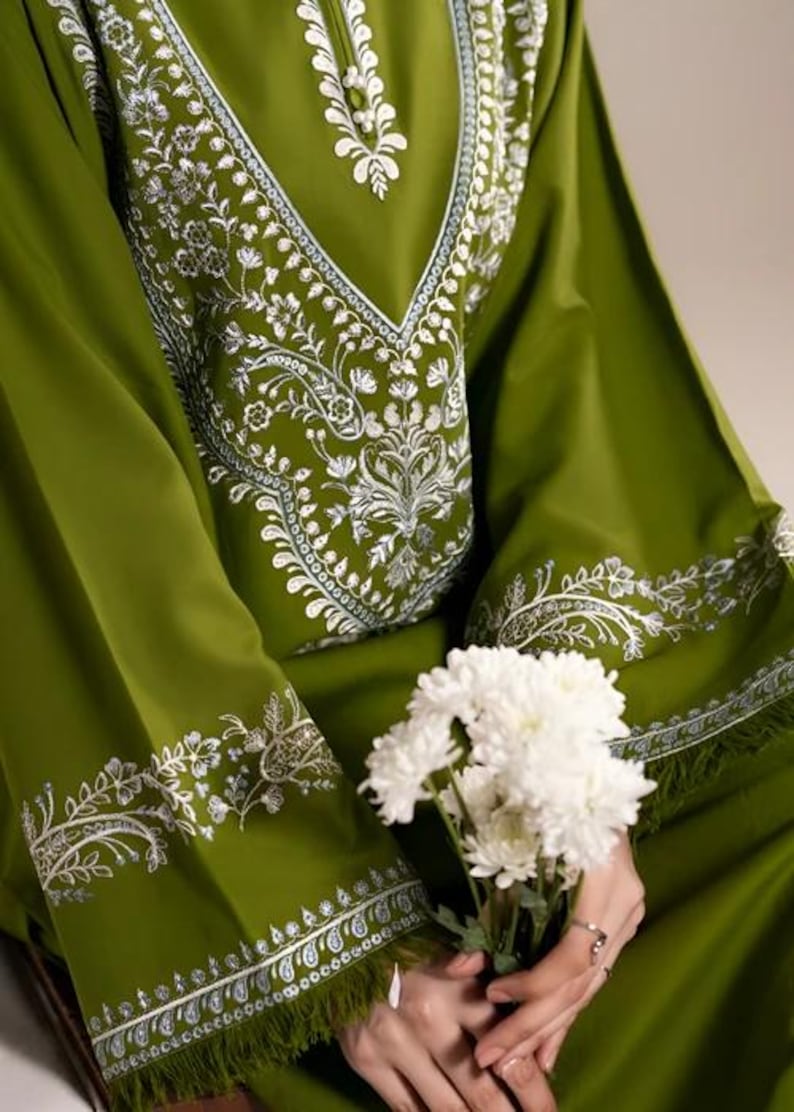 May include: Olive green tunic with intricate white floral embroidery around the neckline and sleeves. The long-sleeved garment features a V-neck design and is paired with a bouquet of white flowers.