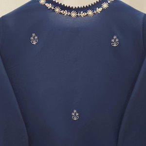 May include: A navy blue top with long sleeves and a round neckline. The neckline is adorned with a delicate floral embroidery in a light gold color. Additional floral embroidery accents are placed on the front of the top.