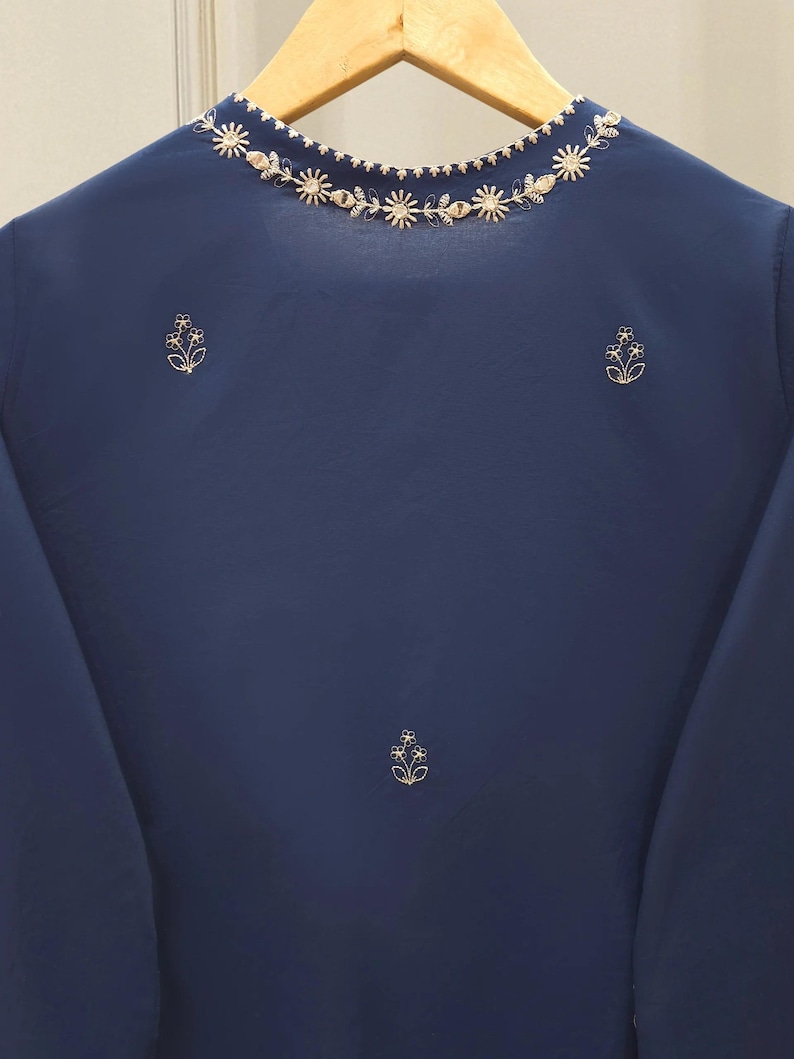 May include: A navy blue top with long sleeves and a round neckline. The neckline is adorned with a delicate floral embroidery in a light gold color. Additional floral embroidery accents are placed on the front of the top.