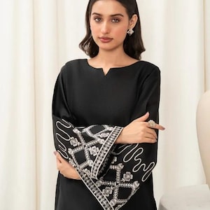 May include: Black long-sleeve tunic with a notched neckline. The sleeves feature a white embroidered design with a geometric and floral pattern. The tunic is a solid black color.