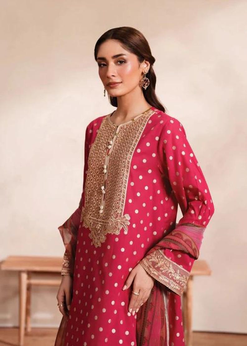 May include: A magenta-colored, long-sleeved tunic with white polka dots. The tunic features an ornate gold embroidered neckline and cuffs, with pearl-like buttons. The sleeves have a patterned border. The outfit is suitable for special occasions.