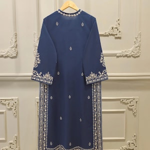 May include: A long, navy blue tunic with intricate white embroidery. The tunic features a round neckline and long sleeves, with detailed floral patterns along the edges and down the front. The garment is hanging on a wooden hanger.