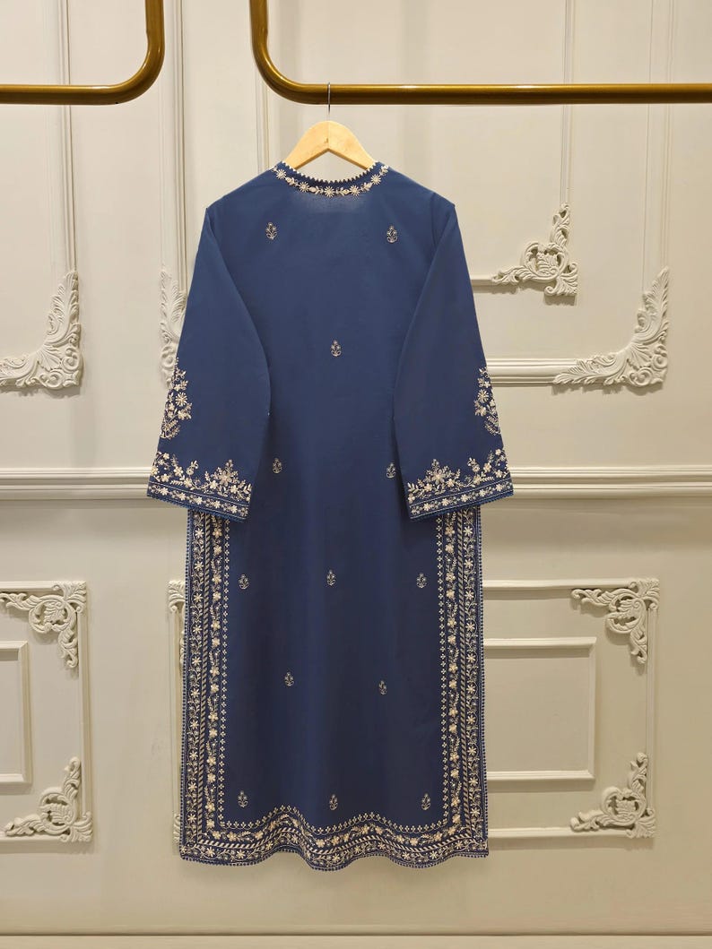 May include: A long, navy blue tunic with intricate white embroidery. The tunic features a round neckline and long sleeves, with detailed floral patterns along the edges and down the front. The garment is hanging on a wooden hanger.